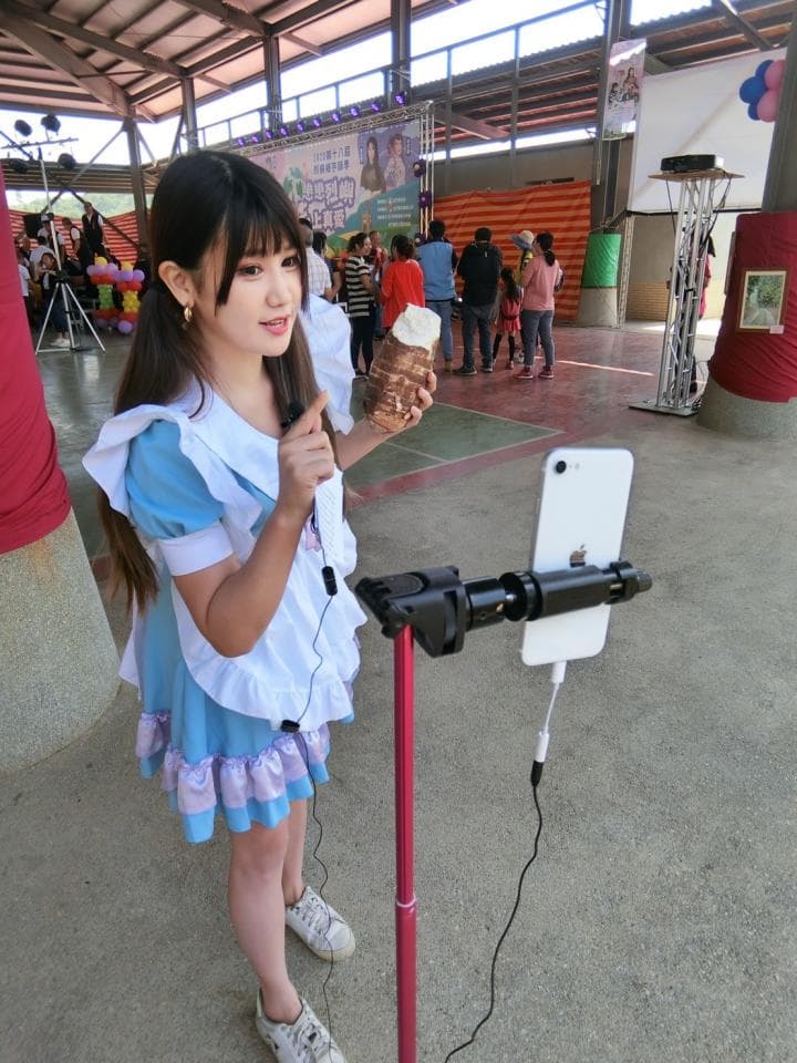 這張「一位穿著女僕裝的女生在錄影」的照片是一個女生穿著藍色女僕裝,手拿著一個冰淇淋,在對著手機錄影,她身後有一個架子,上面放著一個手機,正在錄製她的視頻。