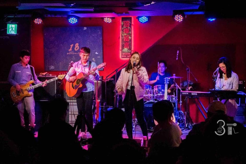 這張「現場演出」的照片是一支樂團在舞臺上表演,其中一位女歌手正在唱歌,一位吉他手正在彈吉他,另外還有一位鼓手和一位貝斯手。舞臺上充滿了燈光,氣氛很熱鬧。