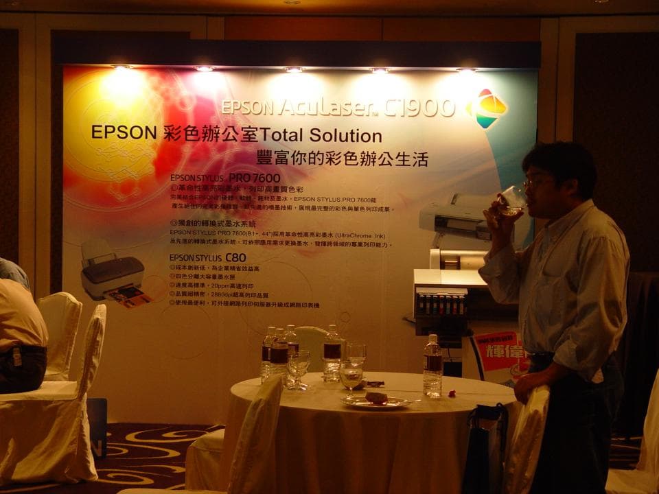 1) 照片顯示一個男人站在一個房間裡,房間裡有一個大的 Epson 廣告,他正在用一個玻璃杯喝東西