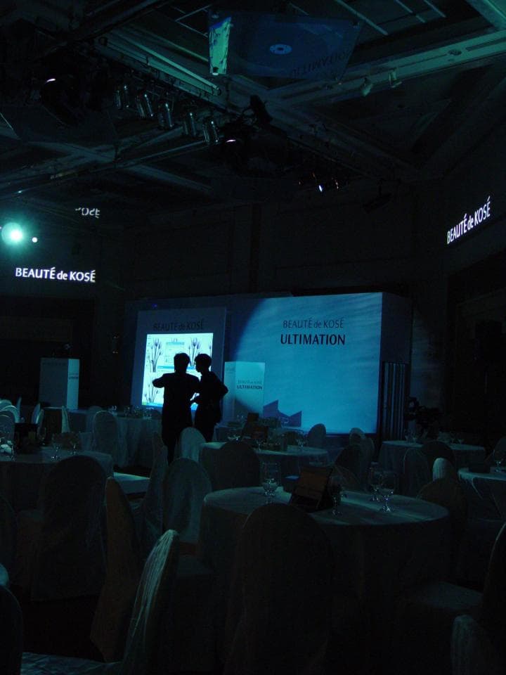 這張照片是一個大型活動的場景,中間的舞臺上有一個大螢幕,上面寫著「BEAUTE DE KOSE ULTIMATION」,周圍擺滿了桌子和椅子,看起來像是正在進行一個產品發布會。