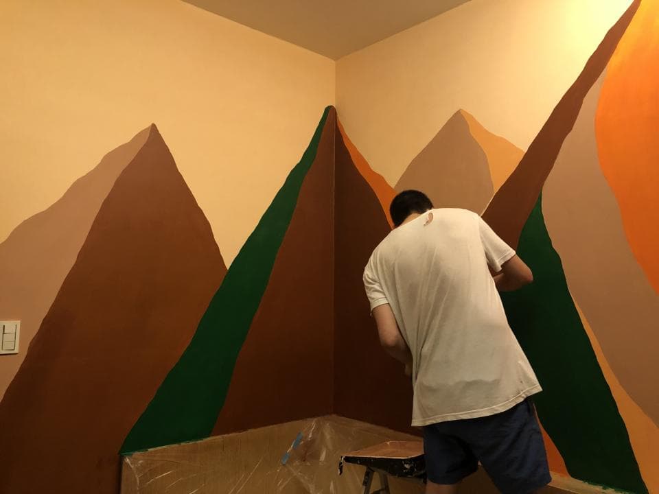 這張照片是一個房間裡正在被粉刷成山景的牆壁。牆壁上畫著以綠色和棕色為主的山脈,一位穿著白色T恤和藍色短褲的男子正在粉刷牆壁。