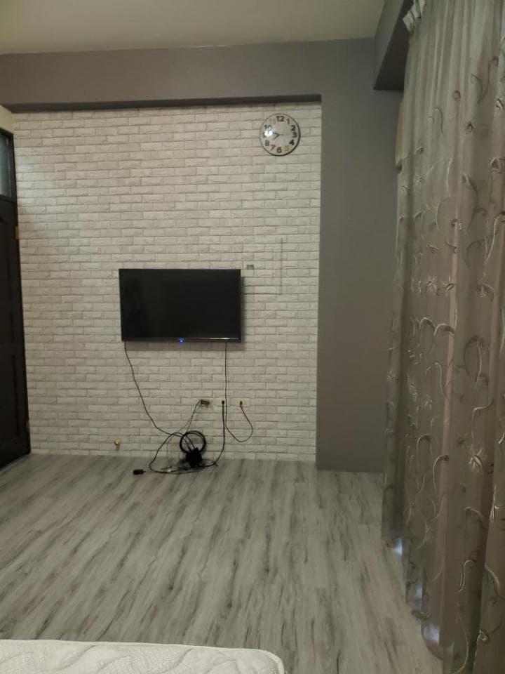 1) 照片顯示一間客廳,牆壁上貼着仿磚牆的壁紙,牆上掛着電視機,房間地面是木紋地板,右邊角落有窗戶,窗戶上掛着窗簾