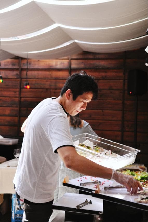 這張「一個男人在餐廳工作」的照片是一個男人正在準備食物,他站在餐桌上,正用他的手從一個透明的容器裡取出食物。他穿著白色襯衫,並戴著白色手套。