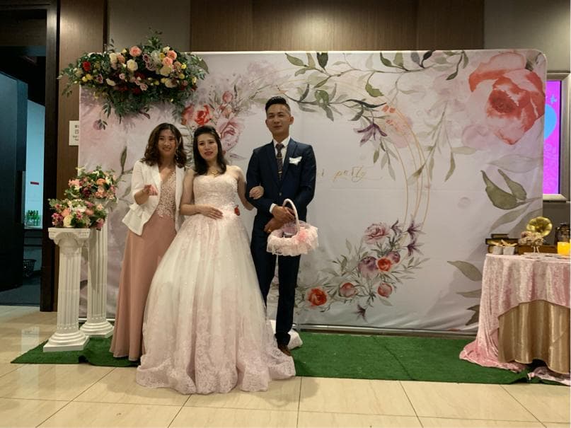 這張照片是一對新人結婚的合照,新娘穿著白色婚紗,新郎穿著藍色西裝,他們站在花牆前,旁邊還有新娘的伴娘。