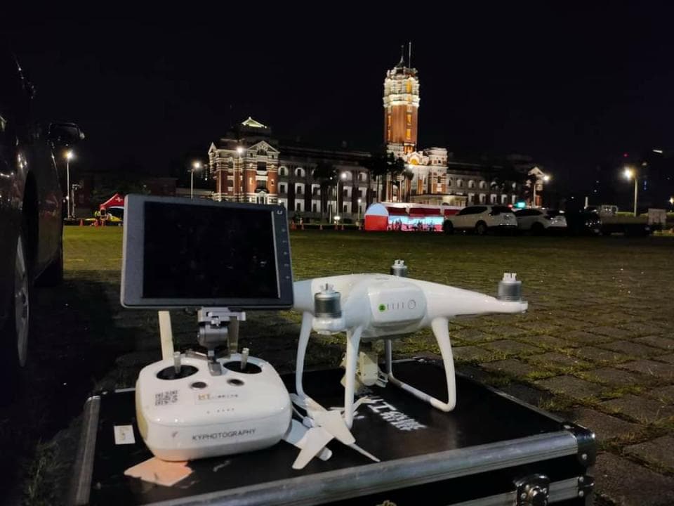 這張照片是一個無人機在晚上被架設在一個黑箱子上,無人機的鏡頭正對著遠處的建築物。無人機旁邊有一個螢幕,可能是用來觀看無人機拍攝的畫面。