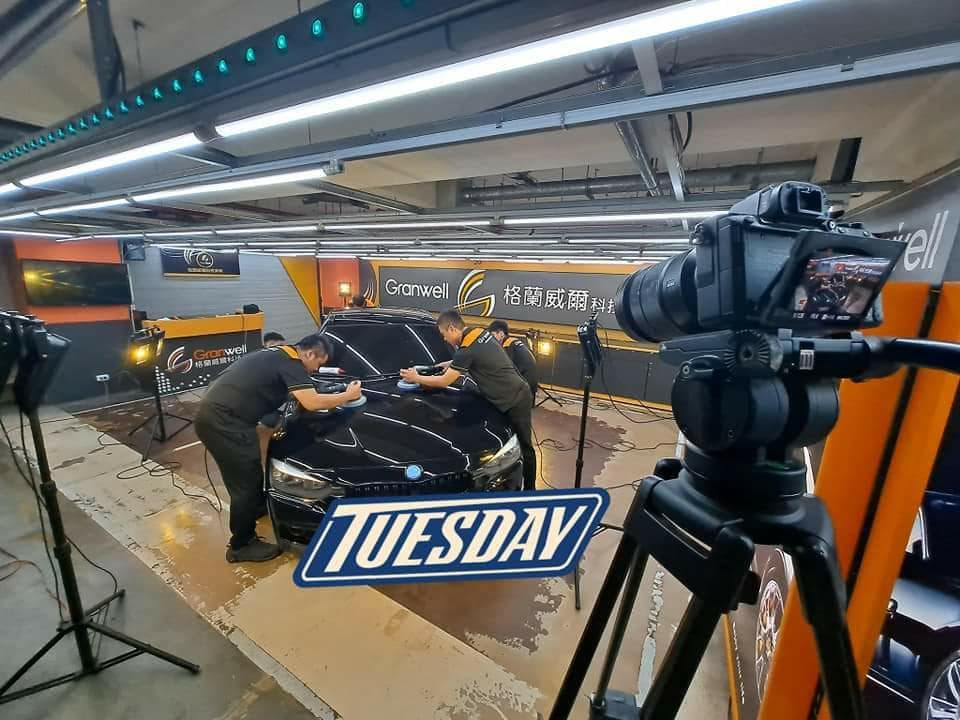 這張「汽車美容」的照片是一個汽車美容店,裡面有工作人員正在用機器為一輛黑色的汽車打蠟,旁邊還有一臺攝影機在拍攝。