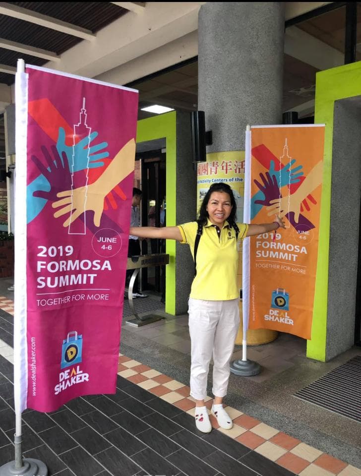 這張「2019 Formosa Summit」的照片是,一個女人站在兩個標語旁,她穿著黃色上衣和白色褲子,笑容燦爛。