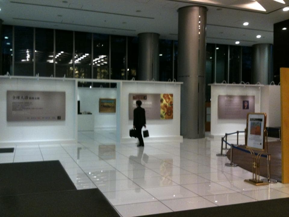 這張照片是一間明亮的展覽空間,空間寬敞,地板光滑,牆壁上掛著幾幅畫作,一位身穿西裝的男子手提公事包,正漫步其中,顯得十分悠閒。