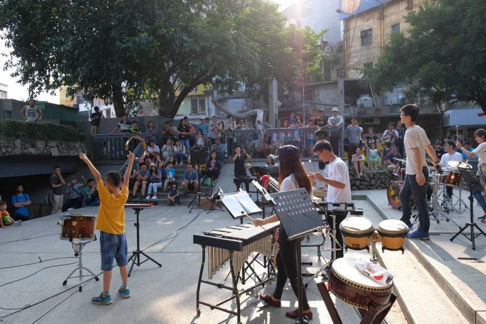 這張「街頭表演」的照片是,一群人在戶外表演音樂。其中有兩位敲打鼓,一位彈奏木琴,還有一位使用手鼓伴奏。他們似乎在一個有建築物的場地表演,周圍有一些觀眾。