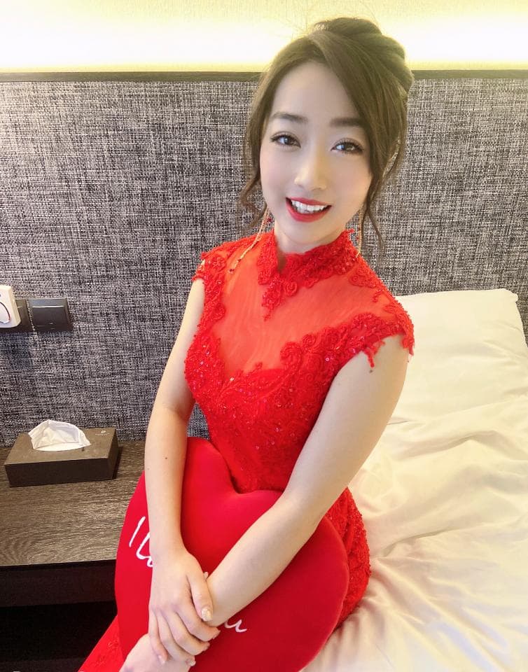 這張照片是一個穿著紅色蕾絲連衣裙的女孩,她坐在一張床上,抱著一個紅色心形抱枕,並露出甜美的微笑。