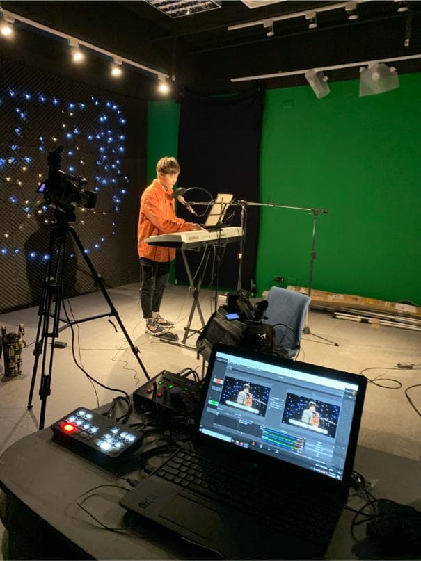 這張照片是一間錄音室,裡面有一個人正在彈奏鍵盤,旁邊放著一個攝像機和一個筆記本電腦,電腦螢幕上顯示著錄製的畫面。