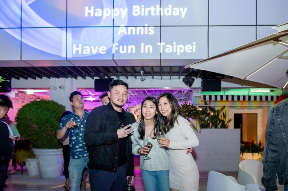 這張「生日派對」的照片是三個人站在一起,他們在一個戶外餐廳,背景上有一個大的招牌寫著「Happy Birthday Ann, Have Fun in Taipei」。