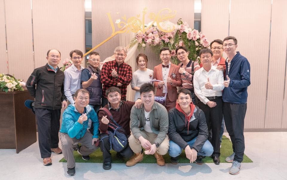 這張「一群人站在花牆前」的照片是,照片中的人們看起來像是朋友或家人,他們都穿著正式的服裝,背景的花牆上裝飾著鮮花,看起來像是在參加婚禮或派對。