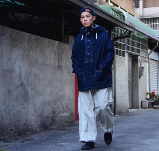 這張照片是一個穿著深藍色外套和白色長褲的年輕男子。他站在一條水泥路旁,背景是幾棟建築物。他看起來很酷,很時尚。