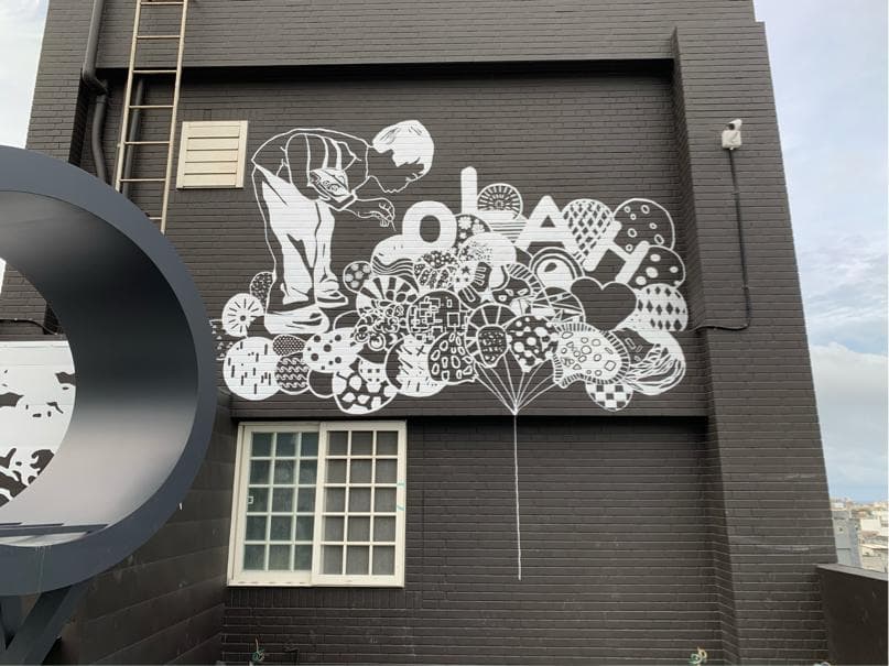 這張「牆壁上塗鴉」的照片是一個黑色的建築物,上面畫著一個白色的人像,周圍是許多白色圓形圖案,像是氣球或泡泡。牆壁上還有白色的字母和圖案,看起來很活潑。