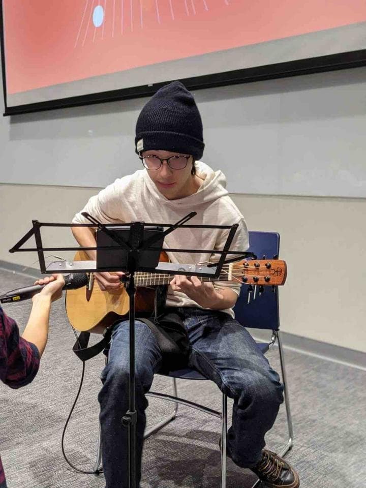 這張照片是一名戴著黑色毛線帽的年輕男子坐在椅子上彈吉他。