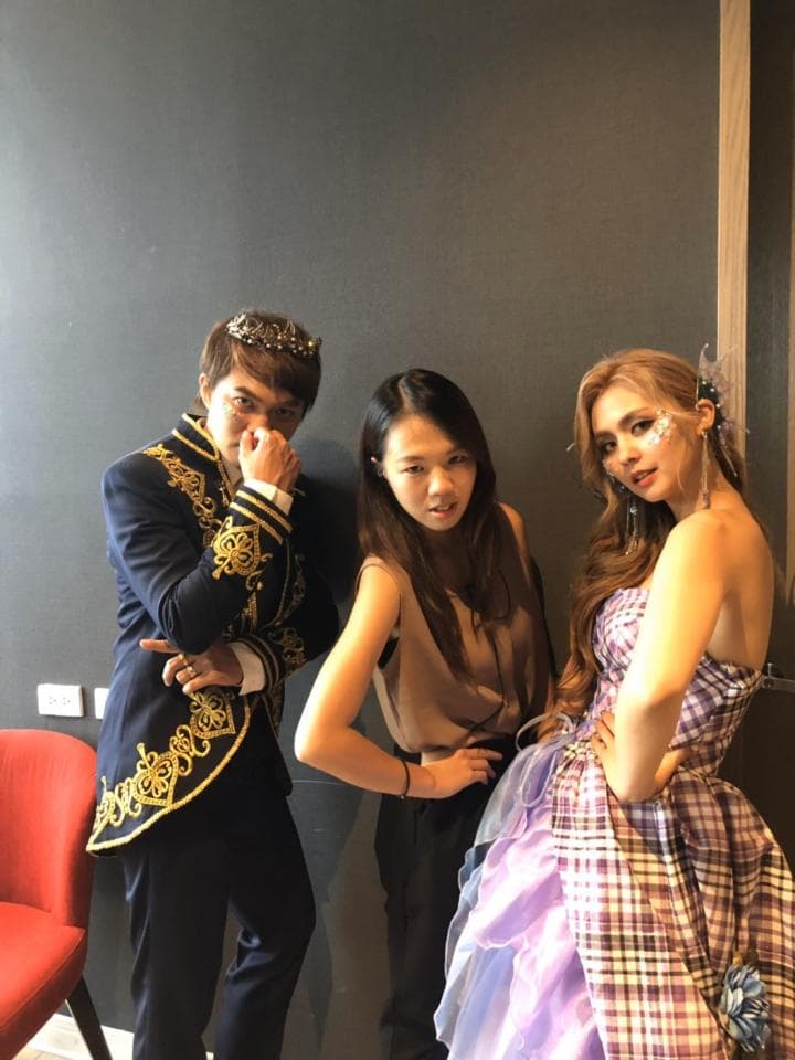 這張照片是三個人站在牆壁前,中間的女孩穿著一件灰色的上衣,左邊的男生穿著一件藍色的外套,上面有金色的裝飾,右邊的女孩穿著一件紫色的連衣裙,上面有格紋。