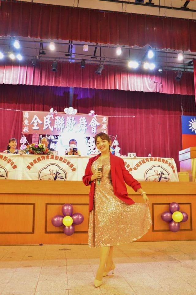 這張「舞臺上表演」的照片是一個女人穿著金色亮片禮服,在臺上拿著麥克風唱歌。她穿著紅色外套,背景是一張桌子和紫色氣球。