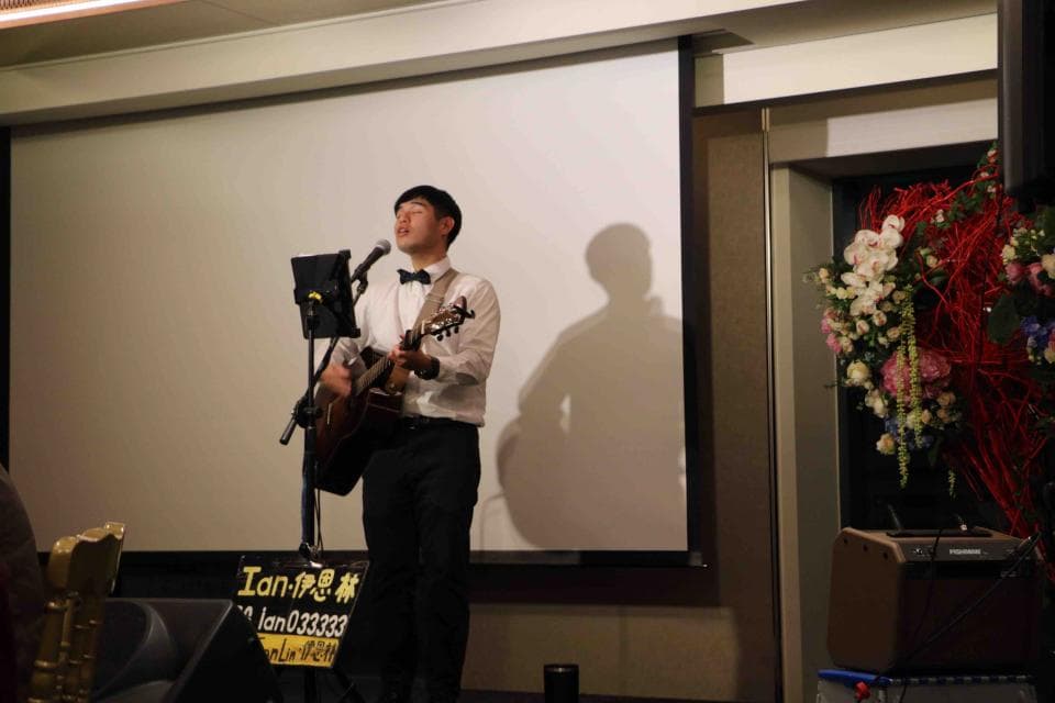 這張照片是一個男人正在舞臺上唱歌彈吉他,他穿著白色襯衫和黑色西裝褲,戴著黑色帽子。他的背後有一個大屏幕,屏幕上有一個影子。