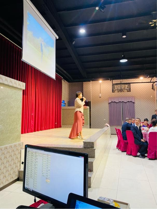 這張「舞臺表演」的照片是一個女性在臺上唱歌,她穿著粉紅色的洋裝,手裡拿著麥克風,臺下有觀眾坐在椅子上,背景有一臺投影機正在播放影片。