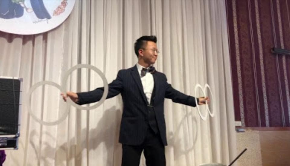 這張「魔術師表演」的照片是一個穿著西裝的魔術師,正在用圓環進行表演,背景是白色窗簾和紅色的牆壁。