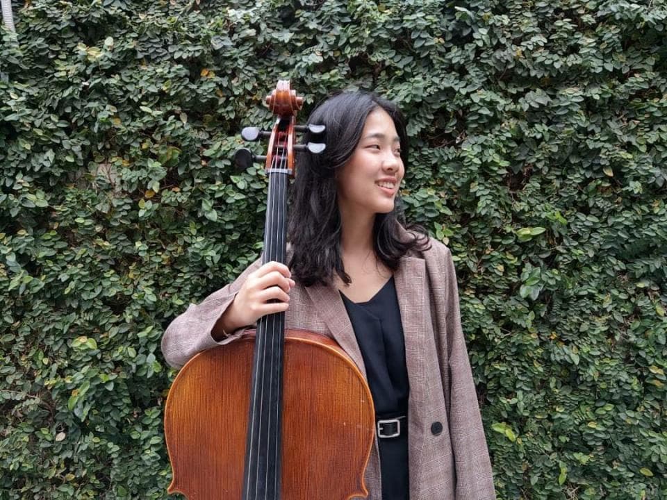 這張照片是一個年輕女子站在綠色植物牆壁前,她抱著一把大提琴。