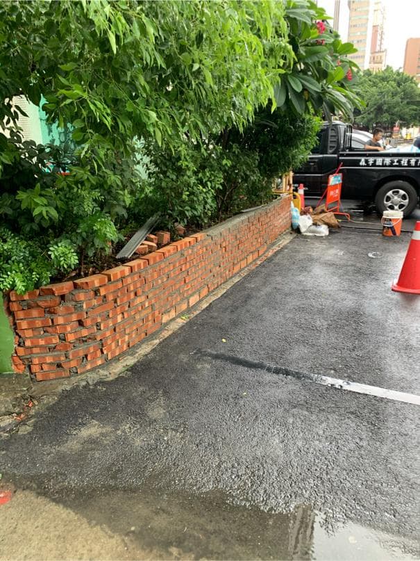 1) 照片顯示一堵磚牆,旁邊是柏油路,左邊是一棵樹,右邊有一輛汽車和一些安全錐