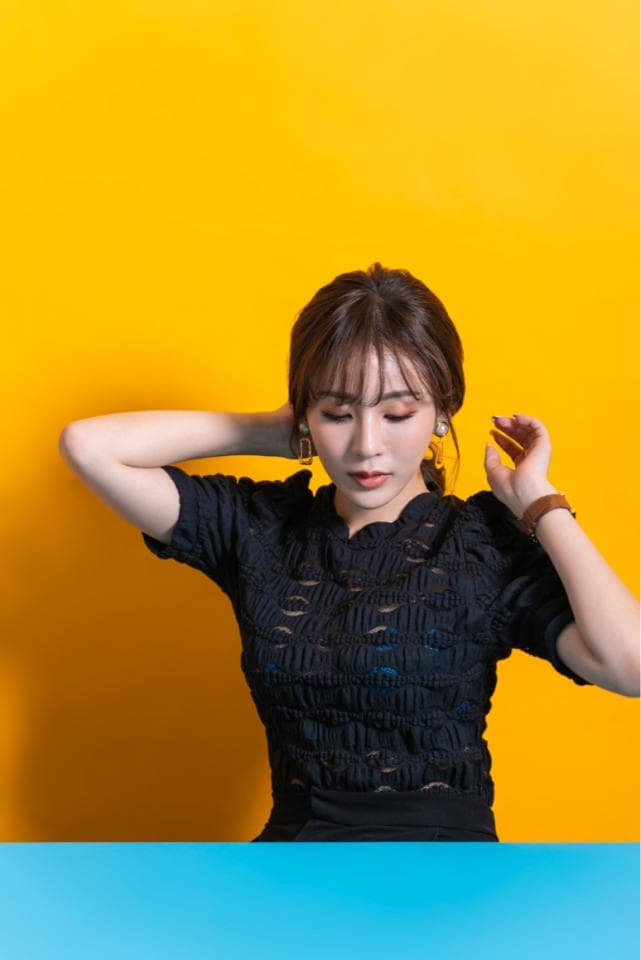 這張照片是一個穿著黑色蕾絲上衣的女孩站在黃色背景前。