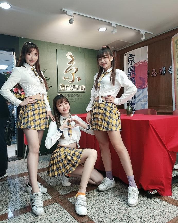 這張照片是三個穿著制服的女生,她們站在一個紅色桌布的桌子上,背景是一面牆,上面寫著「愛爾蘭」和一些其他字。