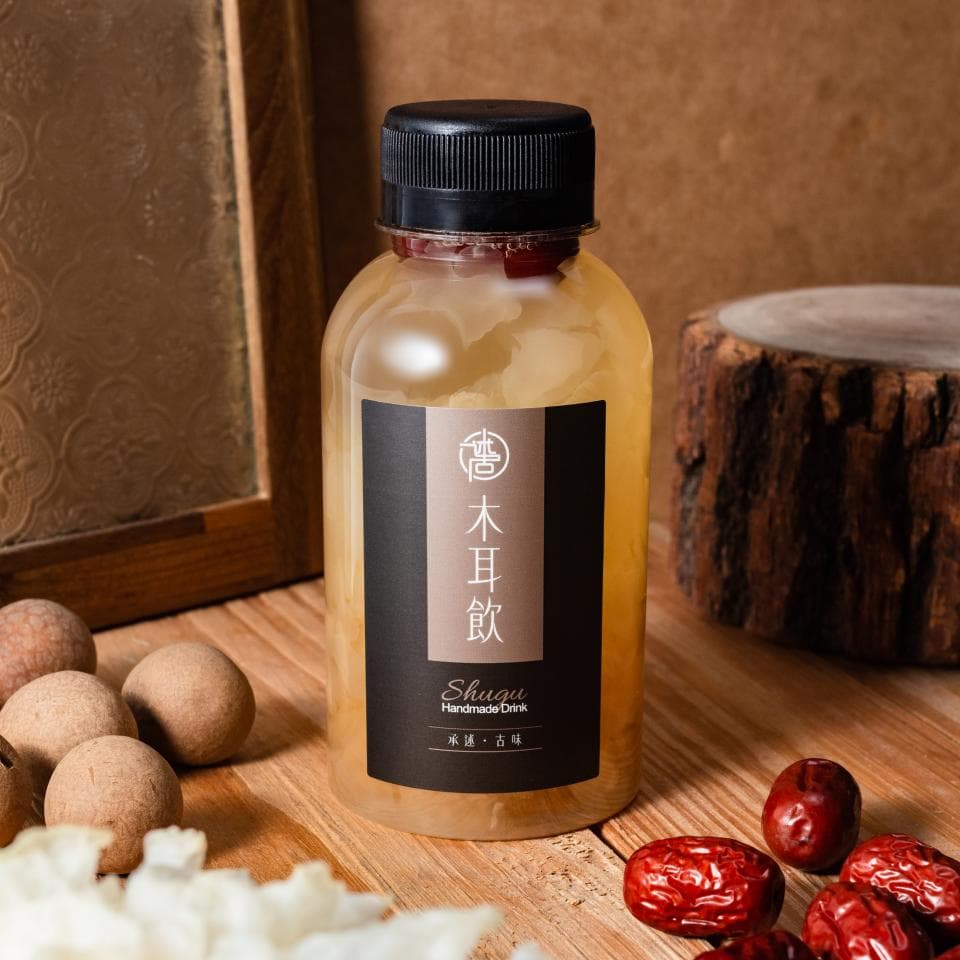 這張「木耳飲」的照片是一瓶裝滿了木耳和金黃色液體的玻璃瓶,瓶子上印著「木耳飲」三個字,還有「Shugu」和「Handmade Drink」的字樣。旁邊有幾顆龍眼和紅棗。