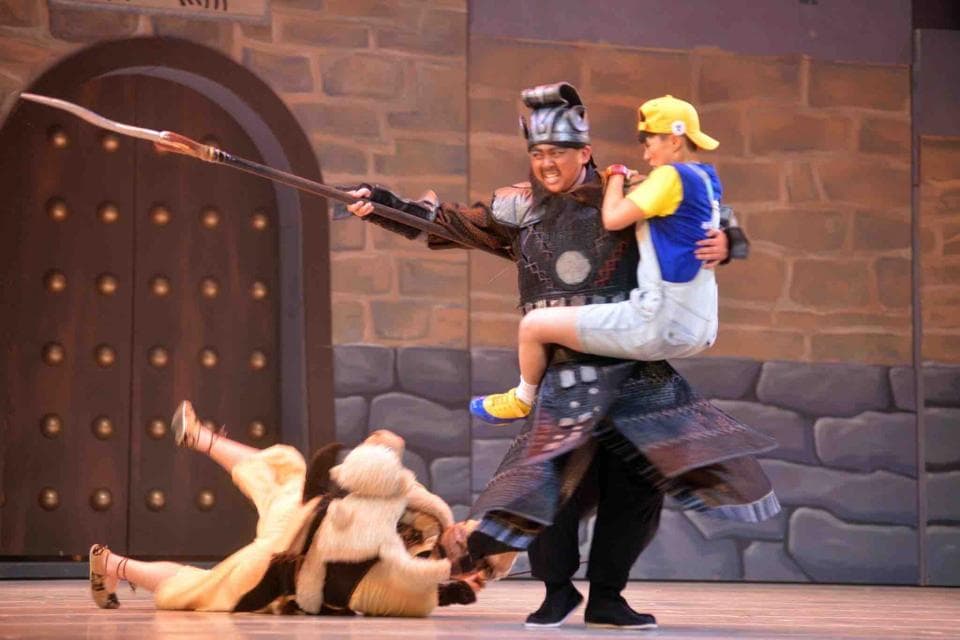 這張照片是一個舞臺劇的演出場景,一位身穿盔甲的將軍正舉起一名身穿黃色上衣的男孩,他手中拿著一根長矛,表情堅定,而男孩則顯得有些驚慌失措。