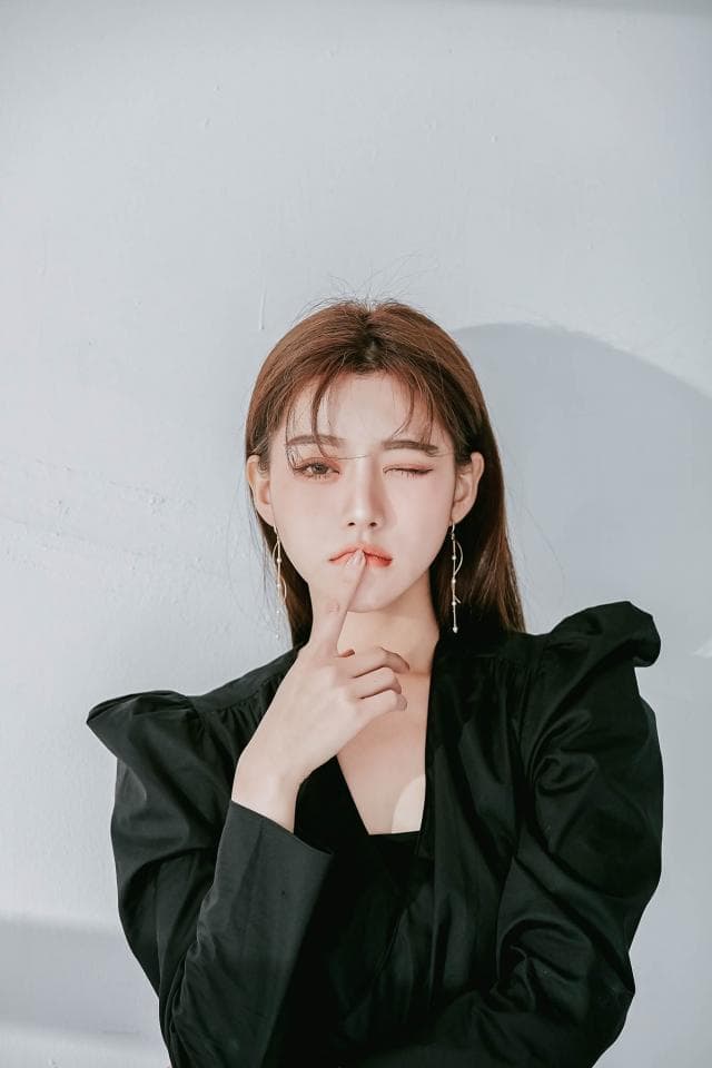 這張「女性肖像」的照片是,一位年輕女性身穿黑色外套,手放在下巴上,看起來很沉思,背景是白色牆壁。