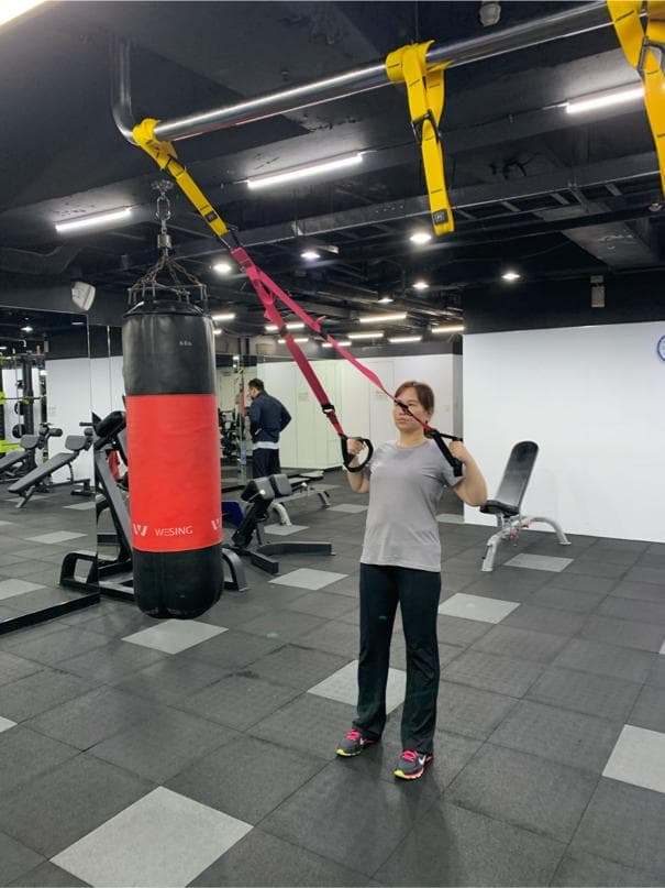 這張照片是一個健身房,最重要的是一位女性正在使用懸吊式訓練器進行鍛鍊。她雙手握住懸吊繩,雙腳微微分開,正在進行上半身的訓練。