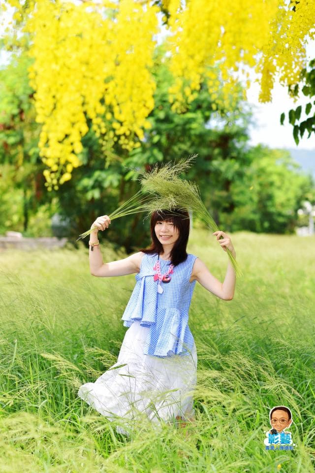 這張「一位年輕女子在金色花樹下,手拿著綠色植物,笑著看鏡頭」的照片是。