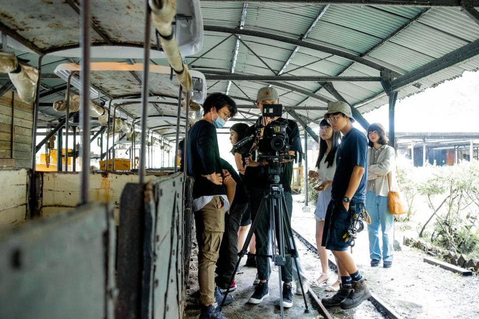 這張照片是一群年輕人在一個廢棄的火車車廂旁拍攝影片。畫面中,一位手持攝影機的男生站在最前面,其他的朋友圍繞在他身旁,似乎正在拍攝一個有趣的片段。