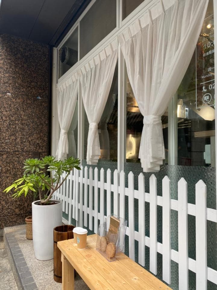 1) 照片顯示一個木質長凳和一個白色柵欄,長凳上放著一個白色馬克杯和一些餅乾,背景是白色窗簾