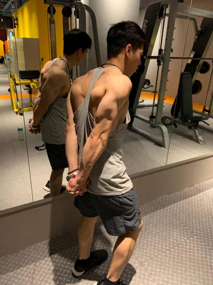 這張「健身房」的照片是一個健壯的男人站在鏡子前,他穿著灰色背心和黑色運動短褲,雙手放在胸前,正在展示他的肌肉。