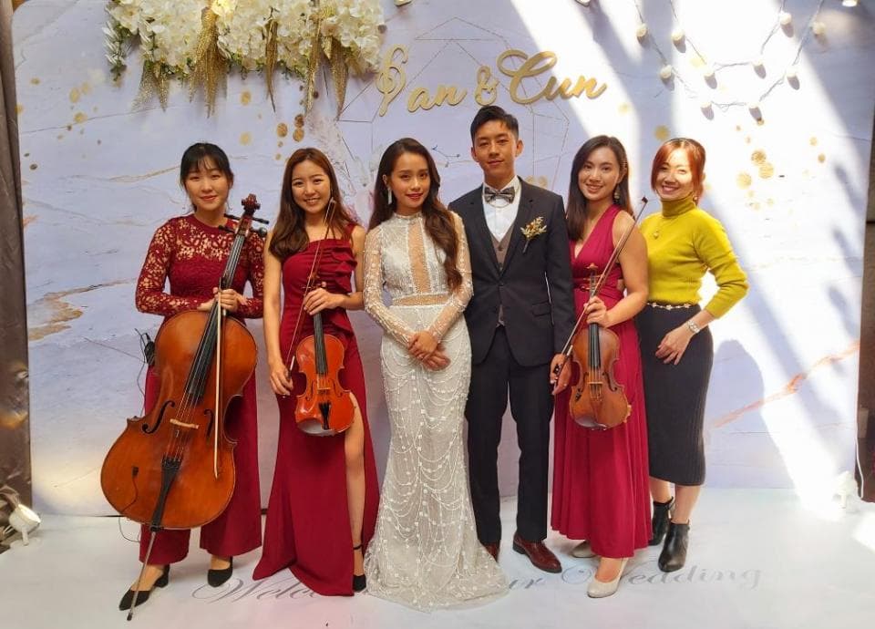 這張婚禮的照片是一對新人在白色背景前,與兩位女樂手和一位男樂手合影。新郎穿著西裝,新娘穿著婚紗,女樂手穿著紅色禮服,男樂手穿著西裝。現場有鮮花裝飾,洋溢著幸福的氛圍。