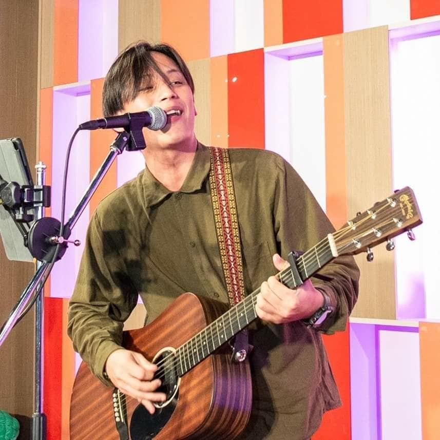 這張照片是一個年輕的男子正在唱歌和彈吉他。他穿著一件綠色的襯衫,並且戴著一條肩帶。他正在用一個麥克風唱歌,他的右手正在彈奏一把木吉他。