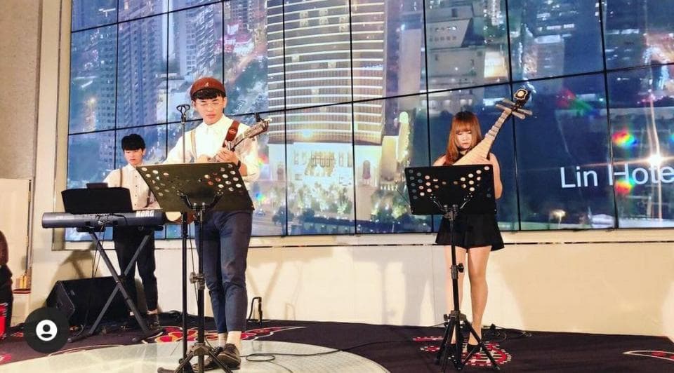 這張「街頭表演」的照片是三個音樂家在表演。其中一位男生彈吉他,另一位男生彈鍵盤,一位女生彈琵琶。他們身後是城市夜景。