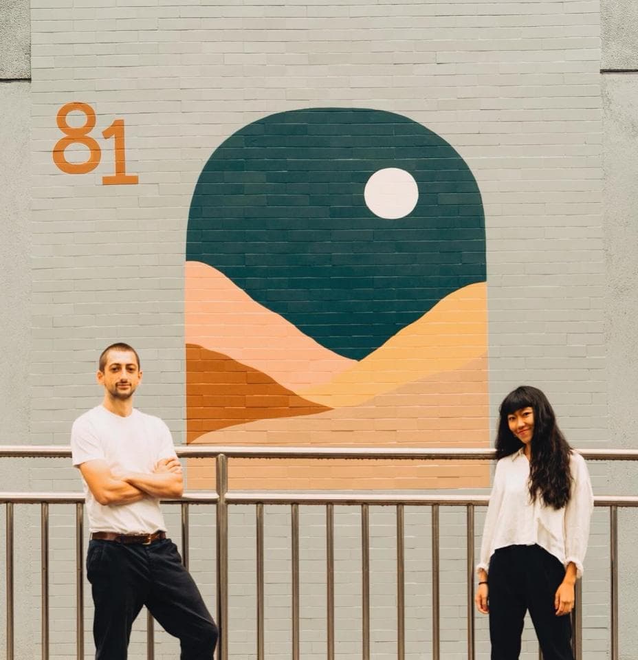 這張「牆壁上有彩繪圖案」的照片是,兩個人站在牆壁前,牆壁上畫著一個拱形,裡面有山丘和月亮。