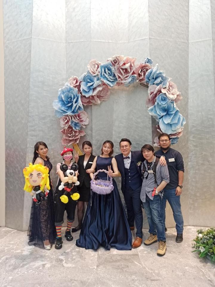 這張「婚禮」的照片是七個朋友在花拱門前拍照,其中一個穿著婚紗,手裡拿著花籃,其他朋友則圍繞在她周圍,有的穿著西裝,有的穿著便服。