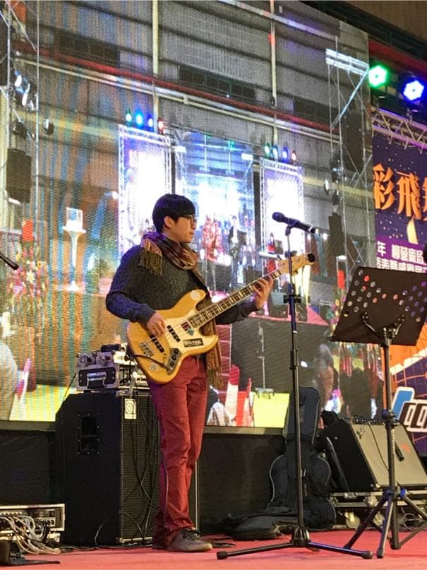 這張「音樂表演」的照片是一個男生在舞臺上彈貝斯,他戴著眼鏡,穿著一件棕色外套,正在演奏。舞臺上放著一個音箱,還有麥克風架。