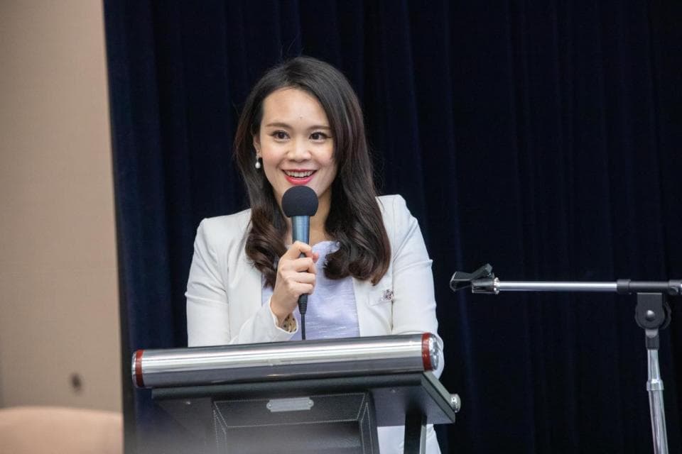 這張照片是一個女人正在演講。她站在講臺上,拿著麥克風,面帶微笑。她穿著白色外套和藍色襯衫,看起來很有自信。