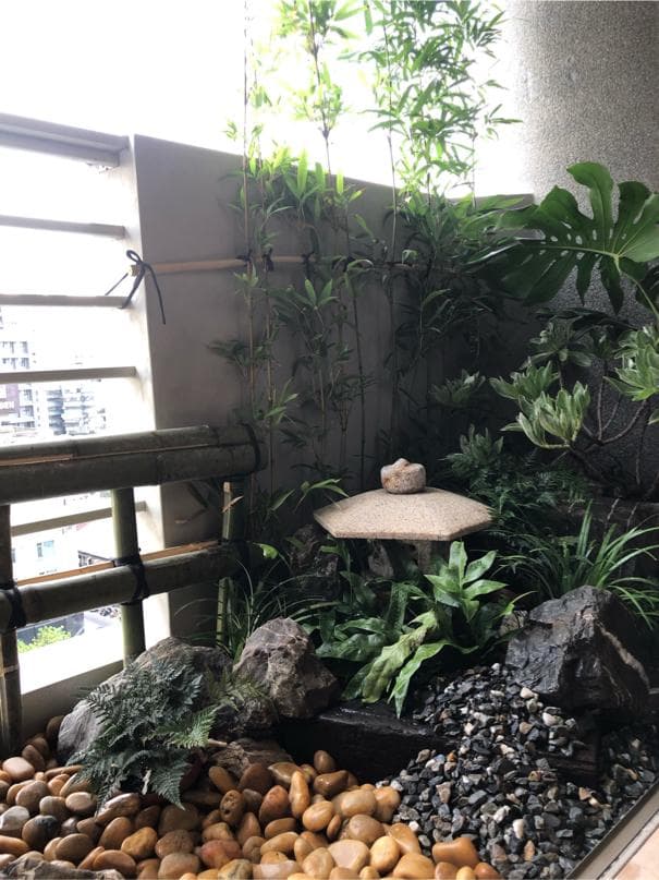 這張「陽臺花園」的照片是一個小型綠色空間,有各種植物和石頭。中間有一座小石燈,旁邊有裝飾石頭。左邊是竹製的欄杆,右邊是水泥牆。整個畫面充滿了自然的氣息。