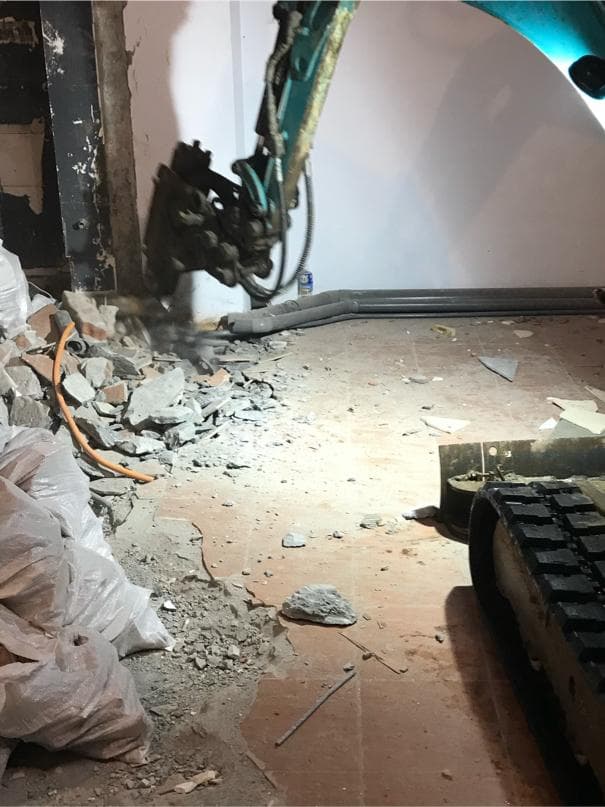 1) 照片顯示一台小型挖土機正在拆除一間房間的牆壁,地面上有許多碎石塊