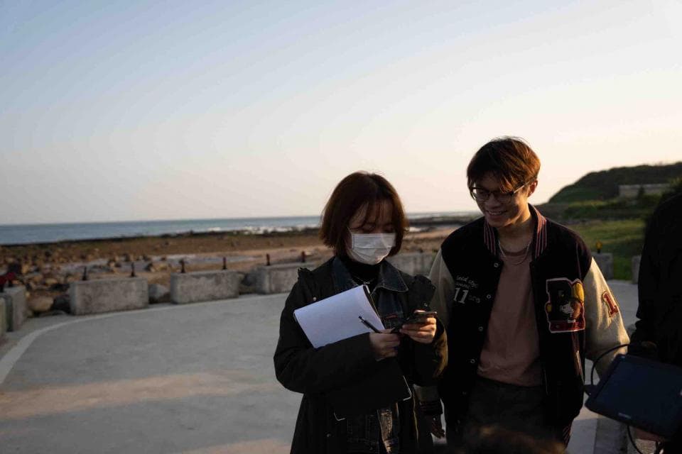 這張「兩個年輕人站在海邊」的照片是,一個戴著口罩的女孩拿著筆記本,另一個戴著眼鏡的男生穿著外套,他們站在海邊的路上。