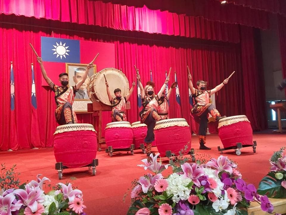 這張照片是五位表演者在舞臺上敲鼓,他們身穿紅色和金色服裝,戴著面具,表情生動。舞臺上擺放著四個紅色大鼓,他們正用鼓棒敲打著鼓面,發出響亮的聲音。觀眾席上擺滿了鮮花,為表演增添了喜慶的氛圍。