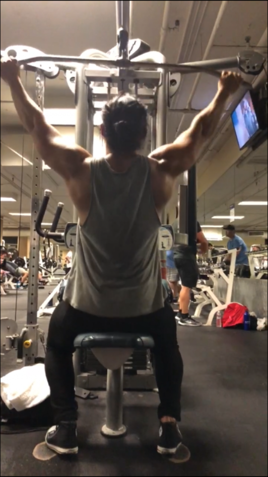 這張照片是一個男子在健身房裡使用拉力器做運動,他背對著鏡頭,雙手抓著拉力器,看起來很努力地做著拉背動作。