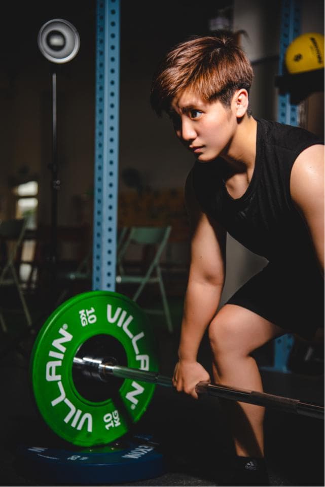 這張照片是一個女性正在舉重。她雙手握著槓鈴,正準備將槓鈴舉起。她的表情專注,身體緊繃,顯示出她正在努力地完成舉重動作。